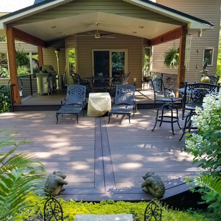 A shaded wooden patio with dark metal lounge chairs and tables, surrounded by lush greenery, ferns, and decorative stone statues.