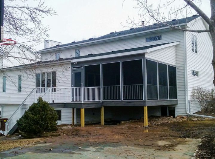 A white-sided home featuring a newly built gray-screened porch with white railings, set against a backdrop of bare trees and a muddy yard.