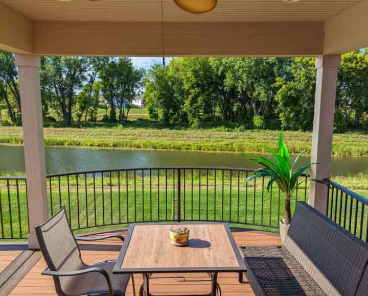 A comfortable wicker patio set on a wooden deck with a view of a tranquil pond and green fields beyond the railing.
