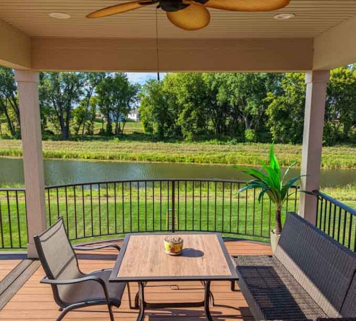 A comfortable wicker patio set on a wooden deck with a view of a tranquil pond and green fields beyond the railing.