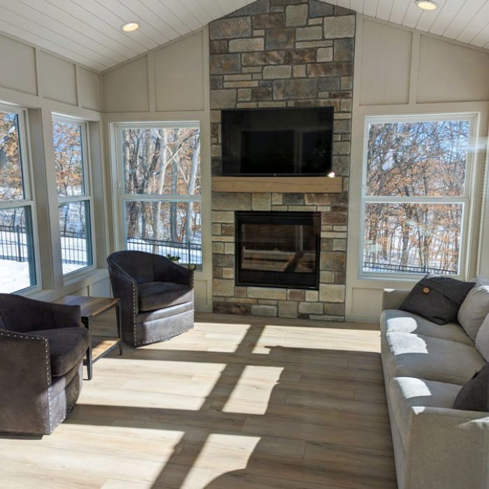 A cozy sunroom featuring a stone fireplace with a mounted TV, light wood floors, white shiplap ceiling, and large windows overlooking a snowy yard.
