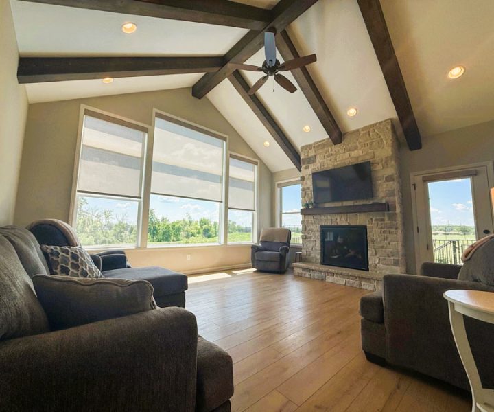 A spacious living room featuring a vaulted ceiling with exposed beams, a stone fireplace, hardwood floors, and large windows offering a view of the outdoors.