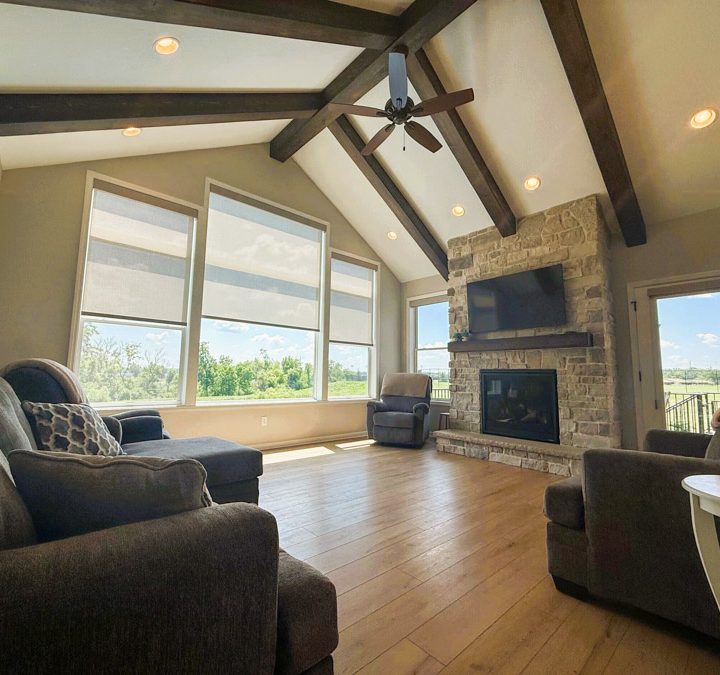 A spacious living room featuring a vaulted ceiling with exposed beams, a stone fireplace, hardwood floors, and large windows offering a view of the outdoors.