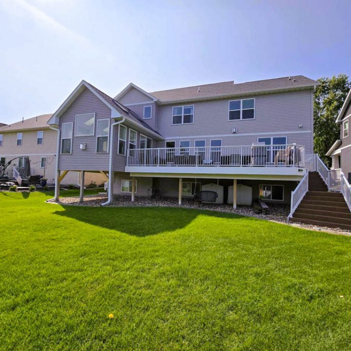 The rear of a light gray home with a white-railed upper deck and stairs, set against a vibrant, expansive green lawn under a clear blue sky.