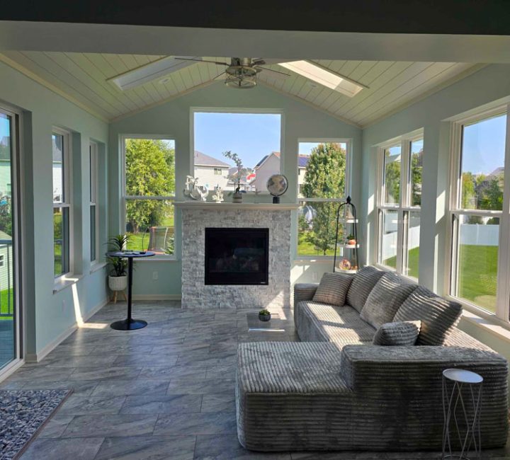 A spacious sunroom featuring a stone fireplace, light blue walls, vaulted ceiling with skylights, and large windows offering views of a green backyard.