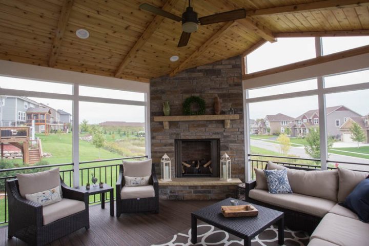A screened porch featuring a stone fireplace, wicker furniture with beige cushions, and large windows offering a view of a green lawn and neighborhood.