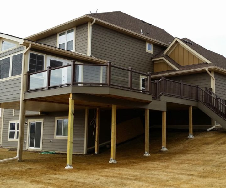 A two-tiered deck with brown railings and glass panels attached to a gray-sided house, situated on a sloped, dry grassy yard.