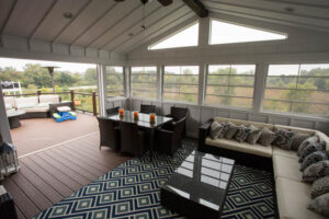 A bright sunroom with an L-shaped beige sofa, a dark dining table, and large windows offering a view of trees and fields outside.