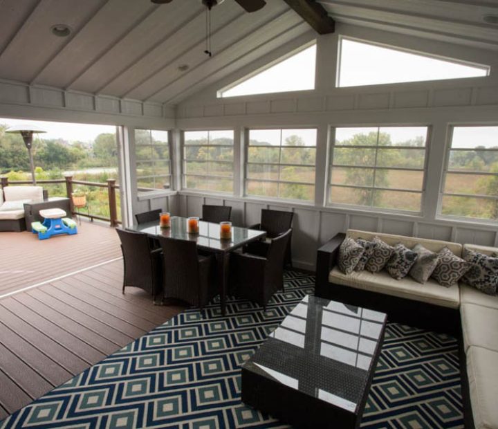 A bright sunroom with an L-shaped beige sofa, a dark dining table, and large windows offering a view of trees and fields outside.