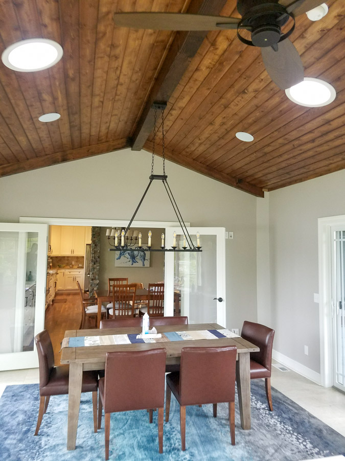 A dining room with a rustic wood ceiling, a large chandelier over a wooden table with brown leather chairs, and an open view into the kitchen.