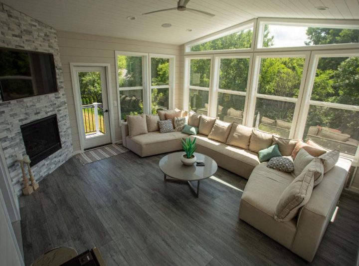 A bright sunroom with an L-shaped beige sofa, a stone fireplace with a mounted TV, and large windows offering a view of lush green trees outside.