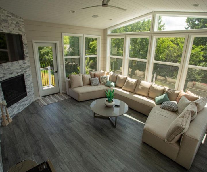 A bright sunroom with an L-shaped beige sofa, a stone fireplace with a mounted TV, and large windows offering a view of lush green trees outside.