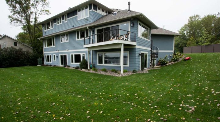 The rear view of a blue two-story house with a lower patio and an upper deck, situated on a sloped green lawn with scattered autumn leaves.