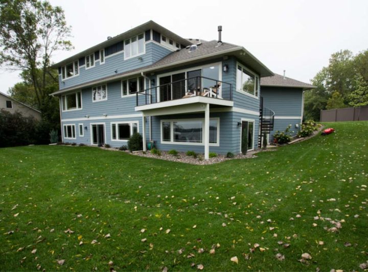 The rear view of a blue two-story house with a lower patio and an upper deck, situated on a sloped green lawn with scattered autumn leaves.