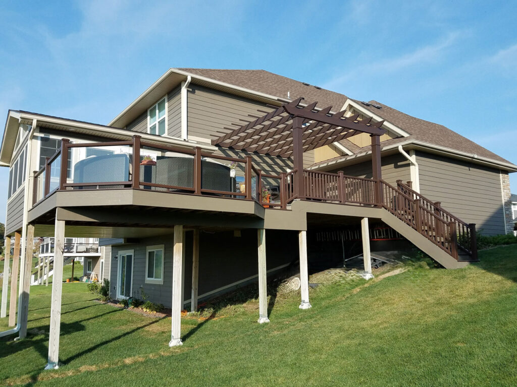 A large, modern deck with brown railings and a pergola attached to a gray-sided house on a sunny day.