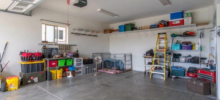 The interior of a clean and organized residential garage with a polished concrete floor. The space is efficiently used with wall-mounted shelving holding colorful storage bins, tool chests, a stepladder, and pet kennels against the far wall.