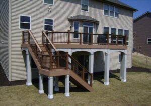 A brown composite deck with matching railings and stairs, supported by white posts, attached to a beige-sided house on a sloped, dry grassy yard.