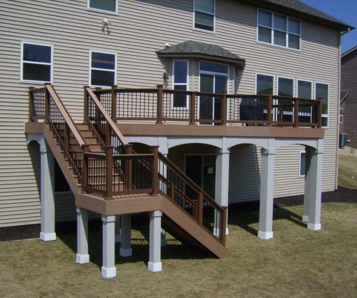A brown composite deck with matching railings and stairs, supported by white posts, attached to a beige-sided house on a sloped, dry grassy yard.