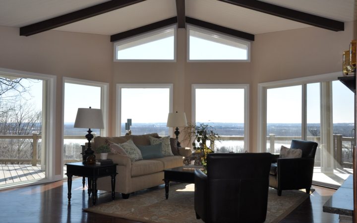 An elegant living area with wide glass windows for natural light to seep in to the interiors, a sofa set and chairs set in a conversational purpose, with carpet, side tables with lamps, and a center table