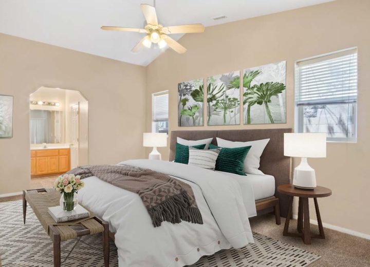 The interior of a bedroom with a carpeted floor, a wide headboard, side tables with lamps, a ceiling fan, wall art display, and own bathroom