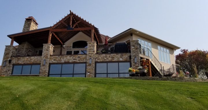 A large, two-story home featuring a stone facade, a prominent chimney, and a spacious covered patio with wooden beams, set against a backdrop of a well-manicured green lawn.