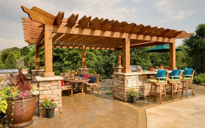 An elegant wooden pergola with stone columns supporting an outdoor kitchen and dining area, complete with bar stools, a table, and vibrant potted plants.