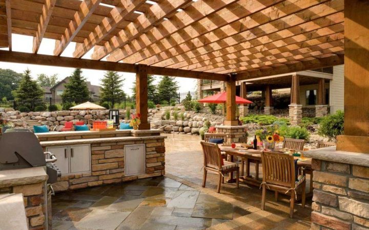An outdoor kitchen under a wooden pergola with stone counters and a built-in grill, set on a paver patio with dining furniture and colorful cushions.