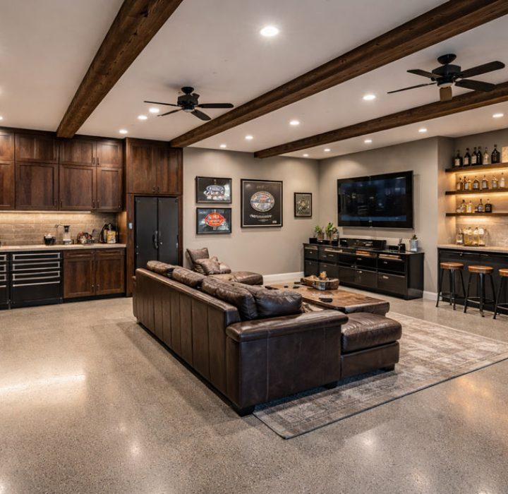 A luxurious finished garage space converted into a man cave or entertainment room. It features polished concrete floors, exposed wood ceiling beams, a leather sectional sofa, a wet bar with illuminated shelving, and custom cabinetry.