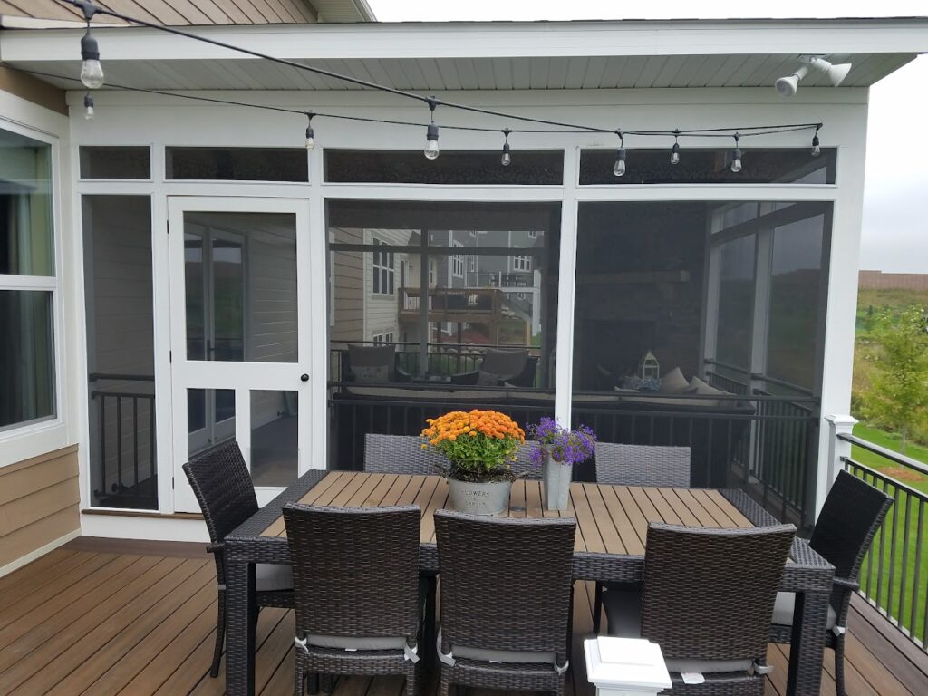 A white-framed screened porch with a dark wicker dining set, vibrant flower centerpiece, and string lights hanging from the ceiling.