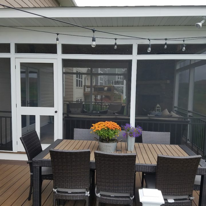 A white-framed screened porch with a dark wicker dining set, vibrant flower centerpiece, and string lights hanging from the ceiling.